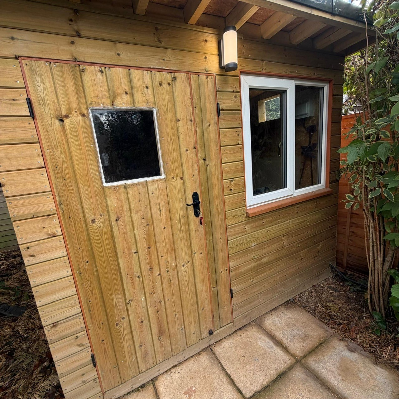 Newly built wooden garden workshop with a lit exterior wall light above the door and a paved patio in front