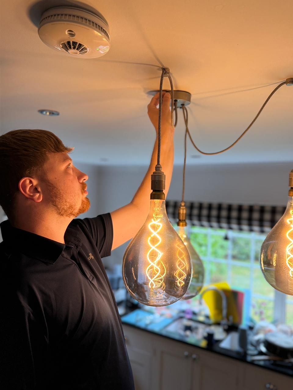 Ollie reaching up to a ceiling mount beside large glass pendant bulbs with warm filament lighting in a kitchen