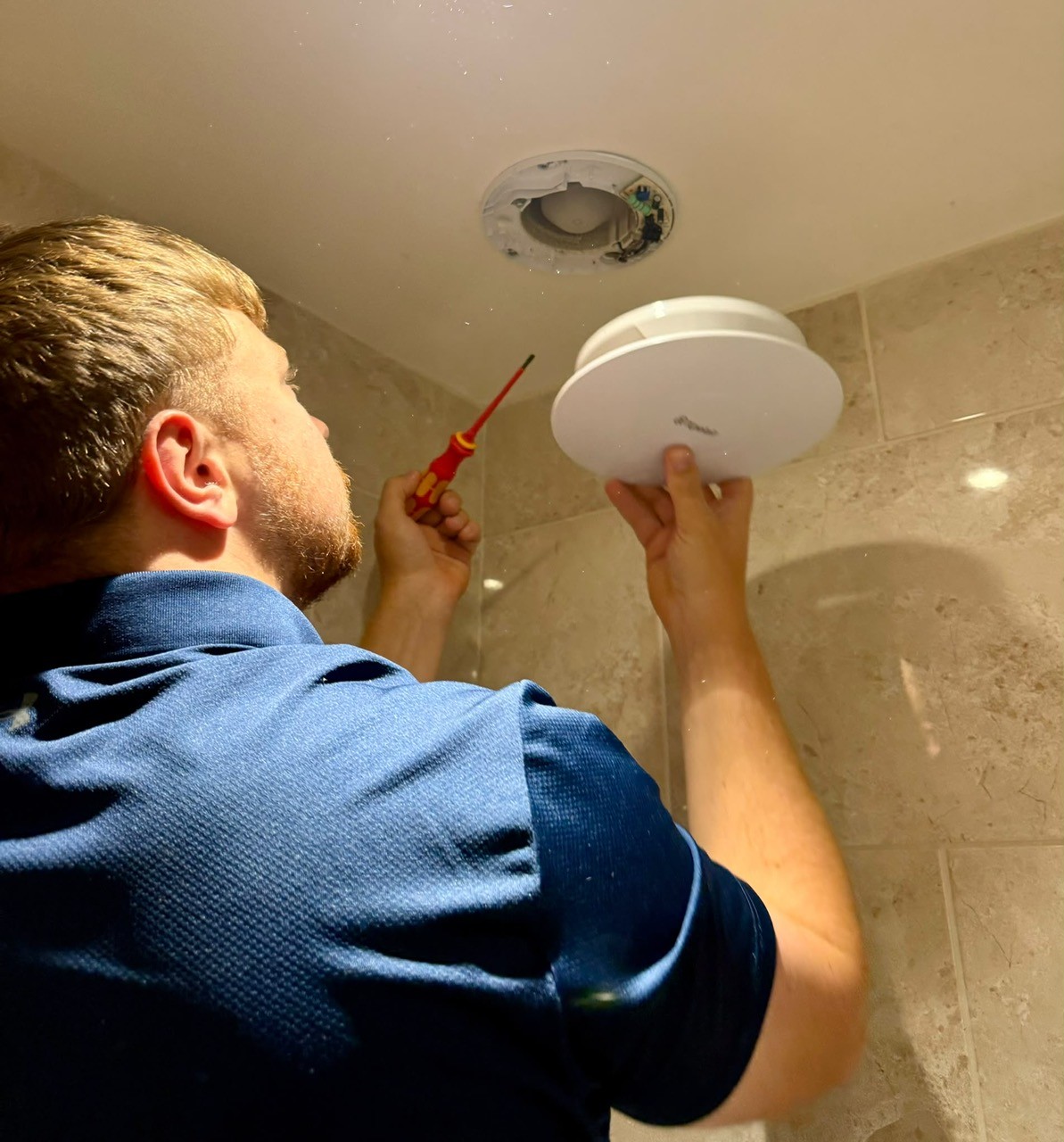 Ollie fitting a round white ceiling extractor fan above a tiled bathroom wall