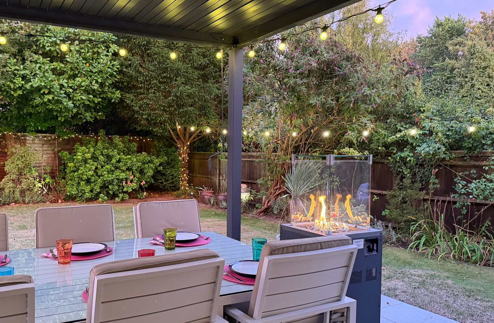 Patio table, fire pit and string lights set up in a garden at dusk